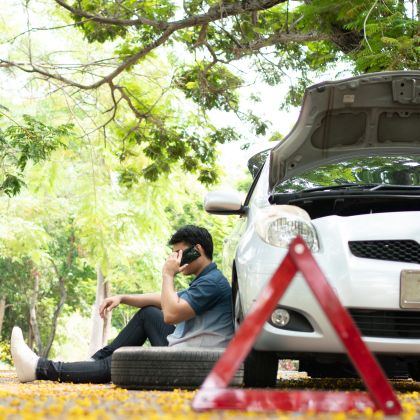 Asian man sitting beside car and using mobile phone calling for assistance after a car breakdown on street. Concept of vehicle engine problem or accident and emergency help from Professional mechanic