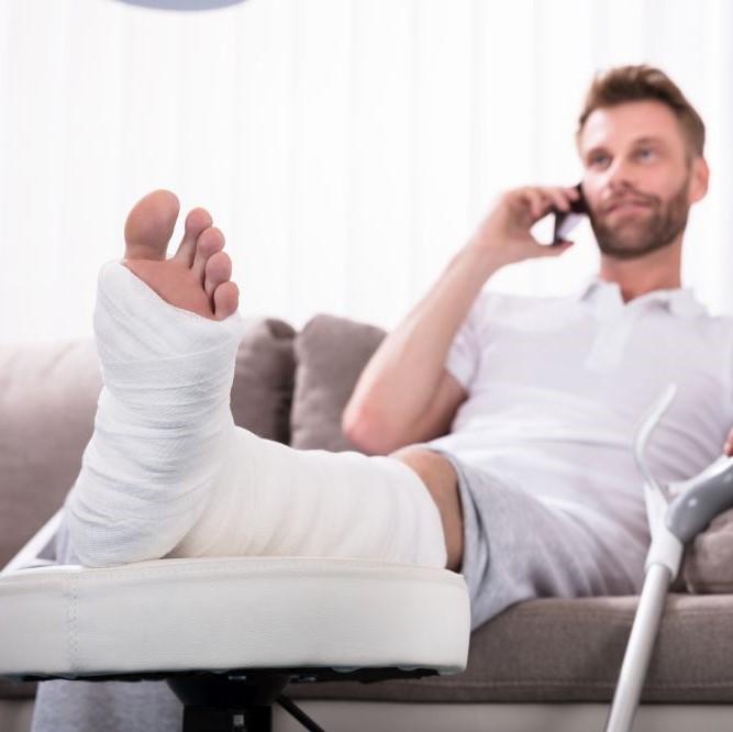 Happy Young Man With Broken Leg Sitting On Sofa Talking On Smartphone