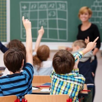 Intelligent group of young school children all raising their hands in the air to answer a question posed by the female teacher, view from behind